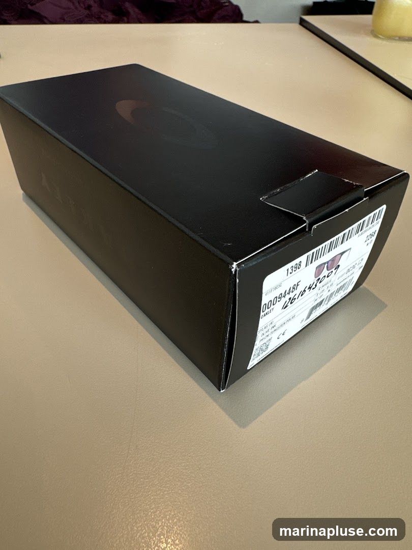 A black Oakley box with a subtle logo on top sits on a light brown surface. A label on the side of the box shows product information, including a barcode and the brand name "OAKLEY".