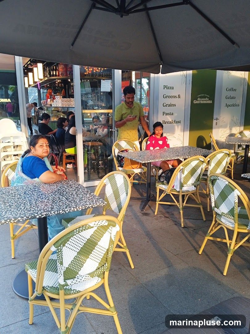 An outdoor seating area of a cafe with tables and chairs. People are seated at some tables, and others are visible inside the cafe through the glass windows. A menu board is visible on the wall with various food and drink options.