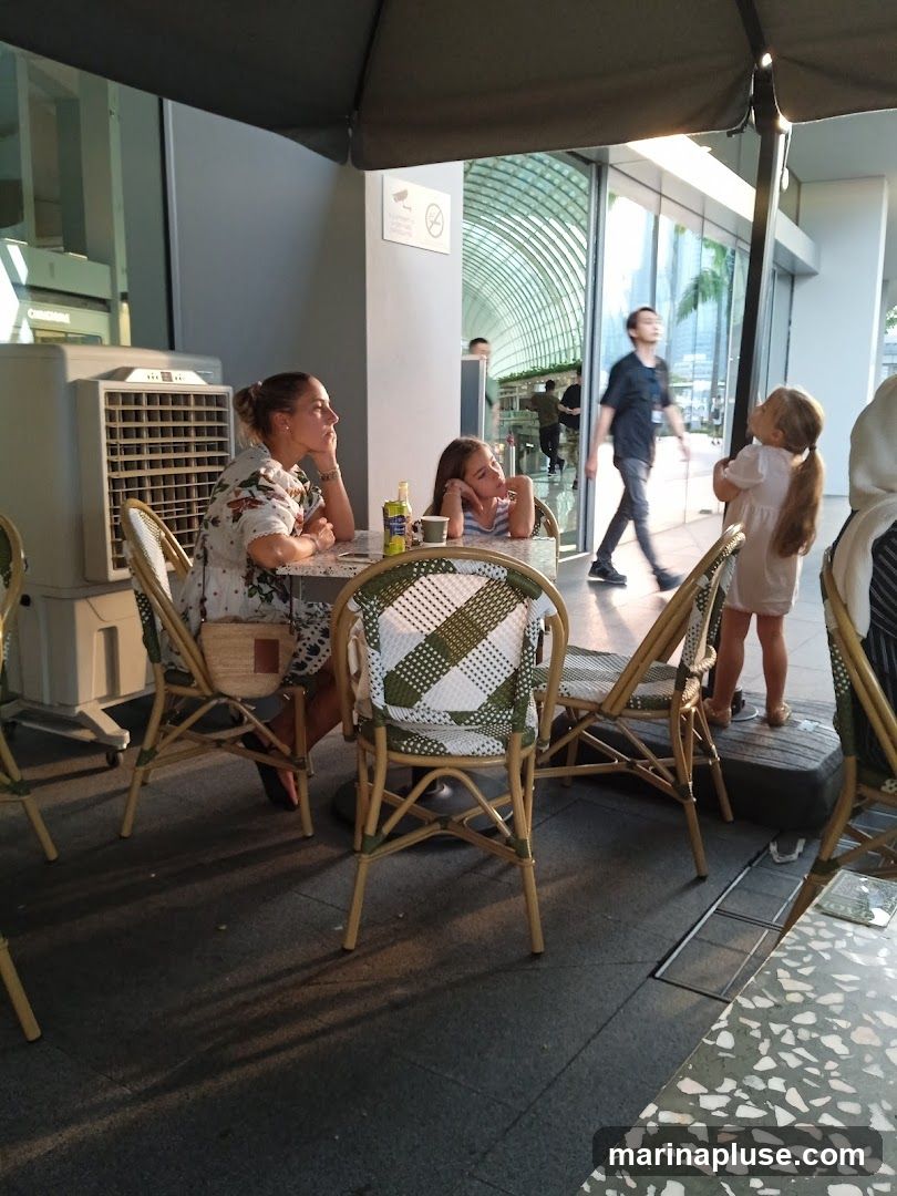 A woman and two children are seated at a table outside a cafe. The woman is wearing a floral dress and the children are looking up. In the background, people are walking past the cafe's glass doors. An air conditioning unit is visible to the left of the table.