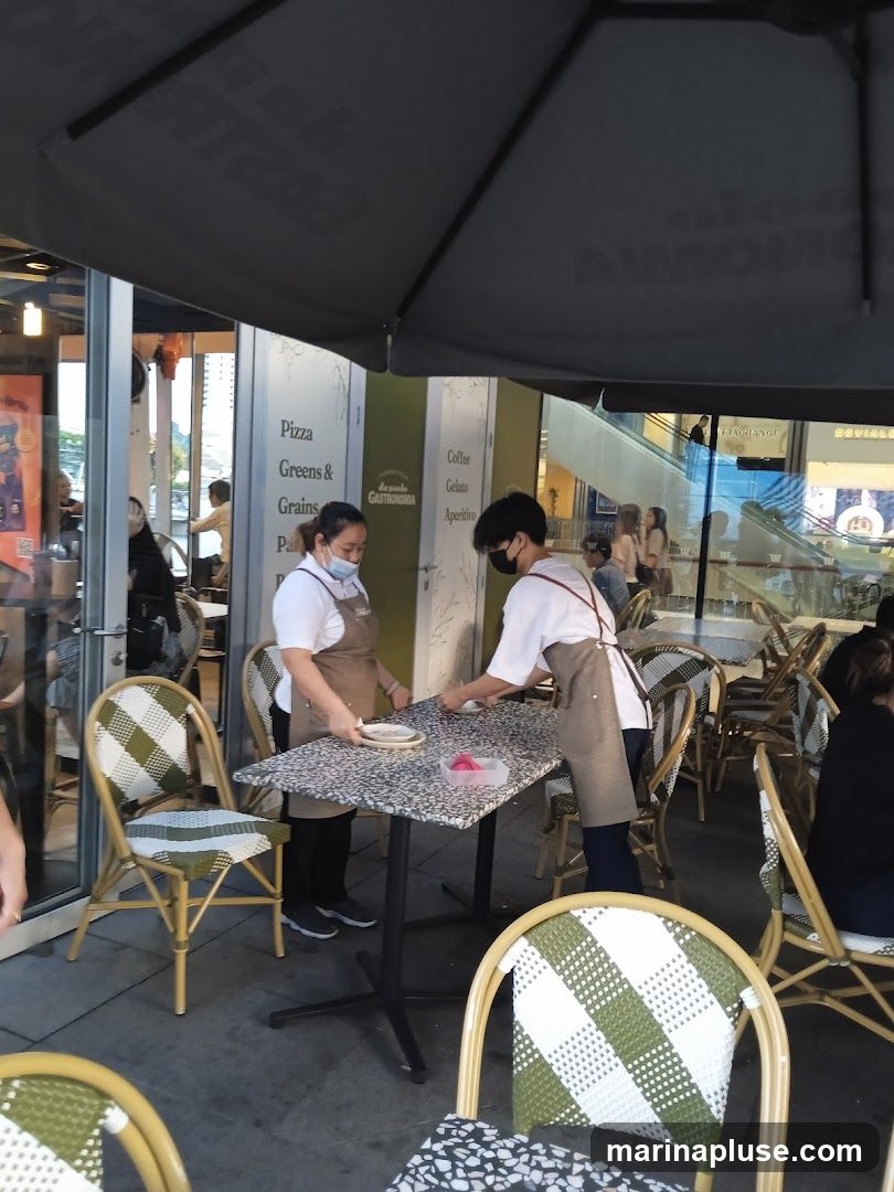 Two servers in aprons are setting up tables outside a cafe. One server is wiping down a table, while the other places items on it. The cafe has outdoor seating with checkered chairs and umbrellas. Signs on the wall indicate offerings like Pizza, Greens & Grains, Coffee, Gelato, and Aperitivo.