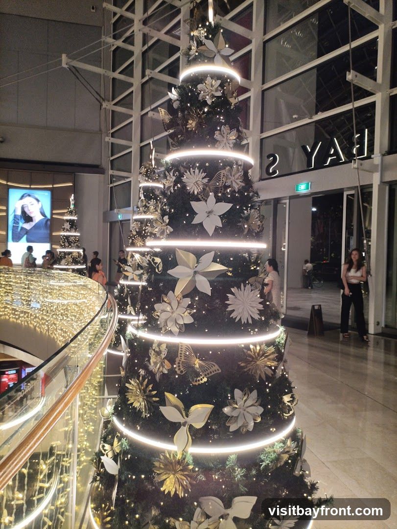 A large, decorated Christmas tree with white and gold ornaments, including flowers and butterflies, is illuminated by strings of lights. In the background, a large digital billboard displays a woman's image, and another smaller Christmas tree is visible. The scene appears to be set in a shopping mall or public space, with people walking around and architectural elements of the building visible.