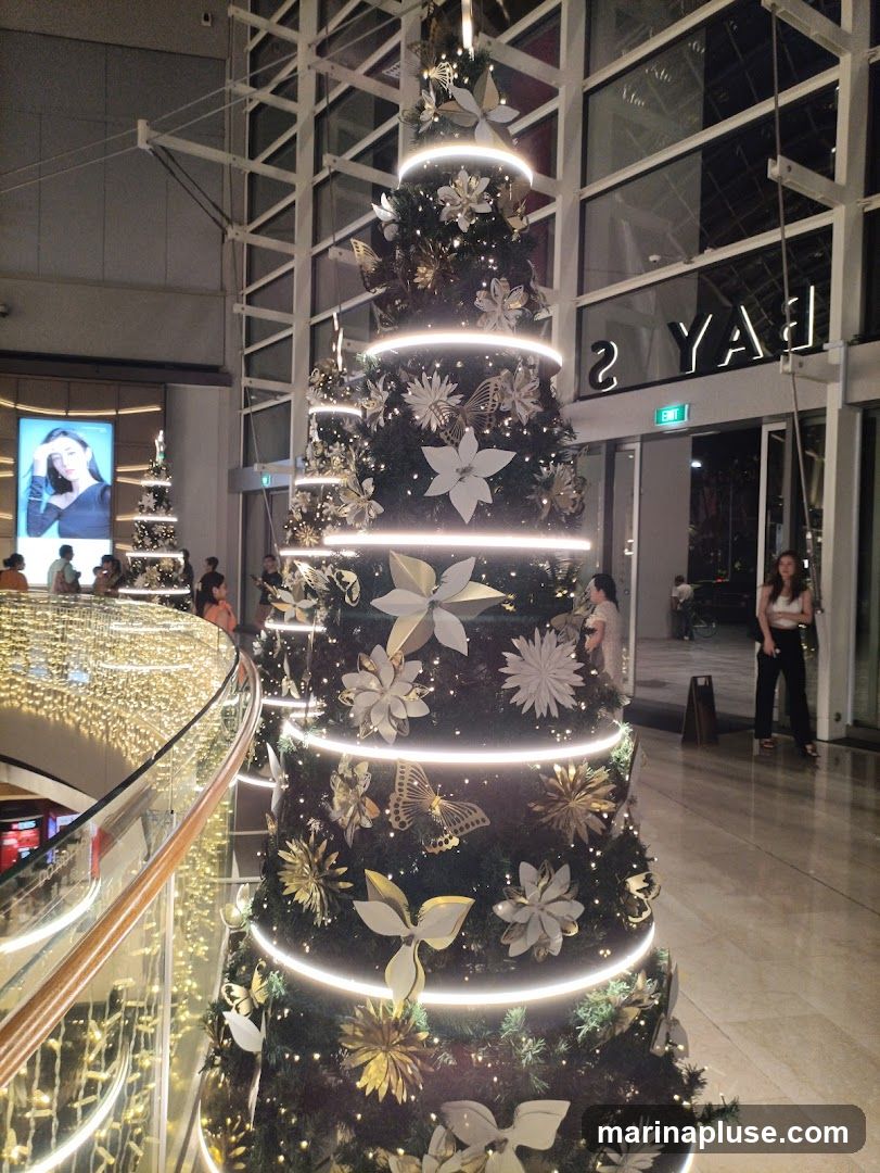 A large, decorated Christmas tree with white and gold ornaments, including flowers and butterflies, is illuminated by strings of lights. In the background, a large digital billboard displays a woman's image, and another smaller Christmas tree is visible. The scene appears to be set in a shopping mall or public space, with people walking around and architectural elements of the building visible.