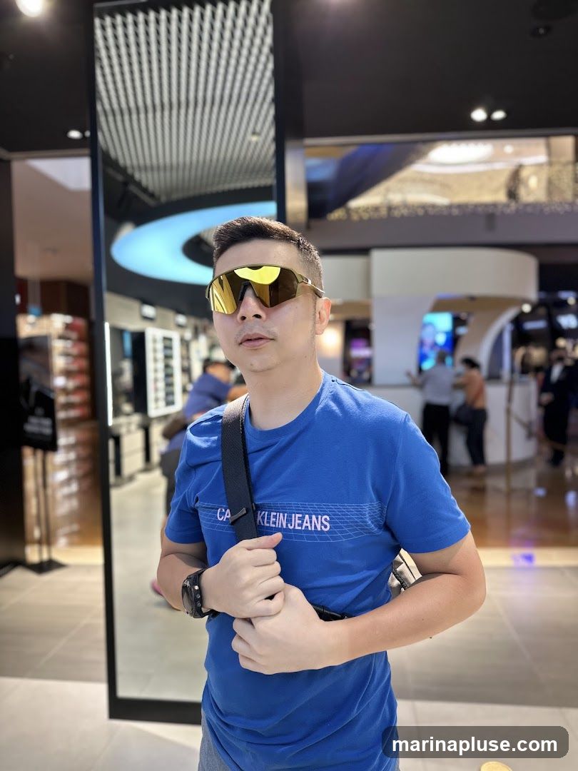 A man wearing large, yellow-tinted sunglasses and a blue t-shirt with "CALVIN KLEIN JEANS" printed on it. He is standing in what appears to be a retail store, with displays of eyewear visible in the background. The store has a modern interior with a large mirror and a curved ceiling light feature. Other shoppers are visible in the distance.