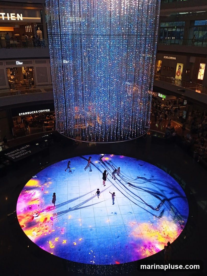 An interior view of a multi-level shopping mall featuring a large, cylindrical art installation made of numerous hanging lights that create a shimmering, blue, starry effect. Below the installation is a circular, interactive digital floor display with projections of colorful abstract art and silhouettes of people walking on it. Surrounding the central area are various shop fronts, including Dior and Starbucks Coffee, visible on different levels.