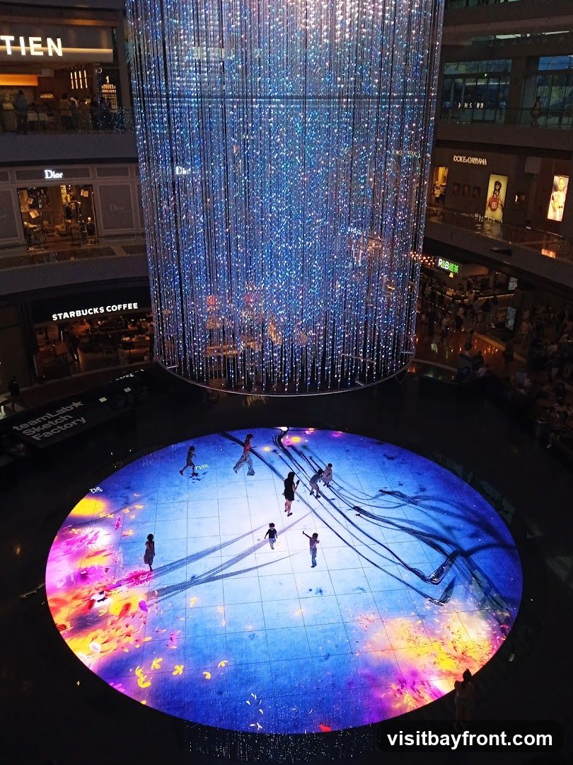 An interior view of a multi-level shopping mall featuring a large, cylindrical art installation made of numerous hanging lights that create a shimmering, blue, starry effect. Below the installation is a circular, interactive digital floor display with projections of colorful abstract art and silhouettes of people walking on it. Surrounding the central area are various shop fronts, including Dior and Starbucks Coffee, visible on different levels.