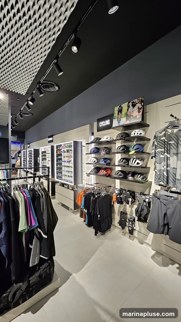 Interior view of a store selling cycling apparel and accessories. Racks of clothing, including jerseys and shorts, are displayed. Shelves are stocked with various cycling helmets. A display case filled with sunglasses is visible in the background. A sign reads "CYCLING" above a display of helmets.