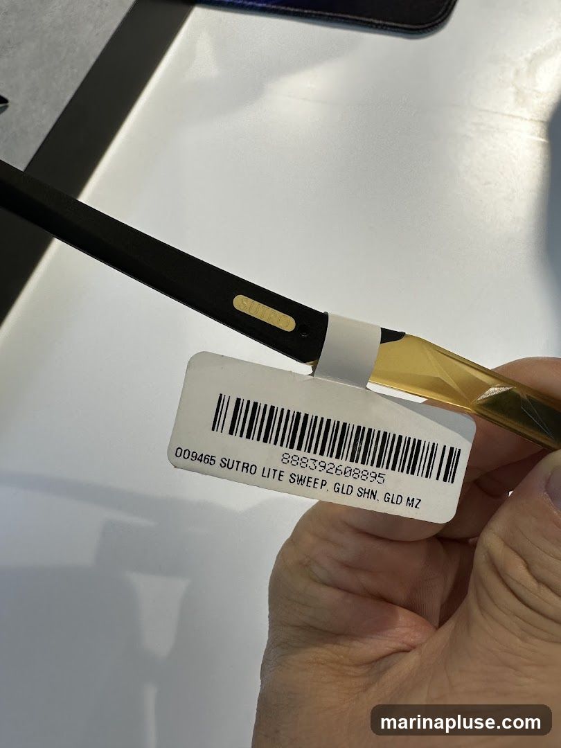 A close-up shot of a product label attached to a black and gold item. The label has a barcode and text that reads "009465 SUTRO LITE SWEEP, GLD SHN. GLD MZ" and a number "888392608895". The item itself appears to be a part of a larger object, possibly eyewear, with the word "SUTRO" embossed on it.