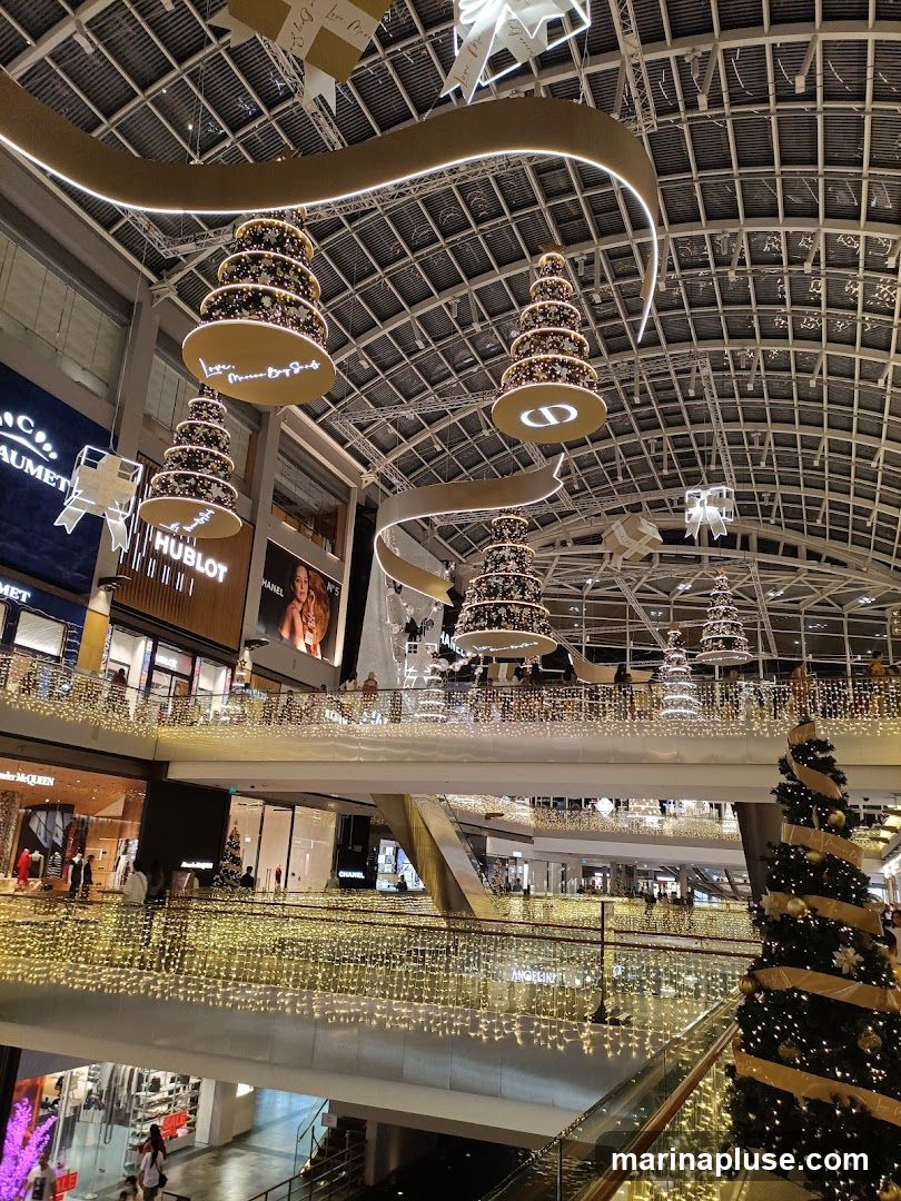 An interior view of a multi-level shopping mall decorated for Christmas. Large, tiered Christmas tree ornaments hang from the ceiling, adorned with lights and gold ribbons. The mall's interior is illuminated by numerous strings of warm white fairy lights draped along the railings and balconies of each level. Storefronts with visible brand names like HUBLOT, CHANEL, and ALEXANDER MCQUEEN are present on the lower levels. A large Christmas tree stands on the right side of the image.