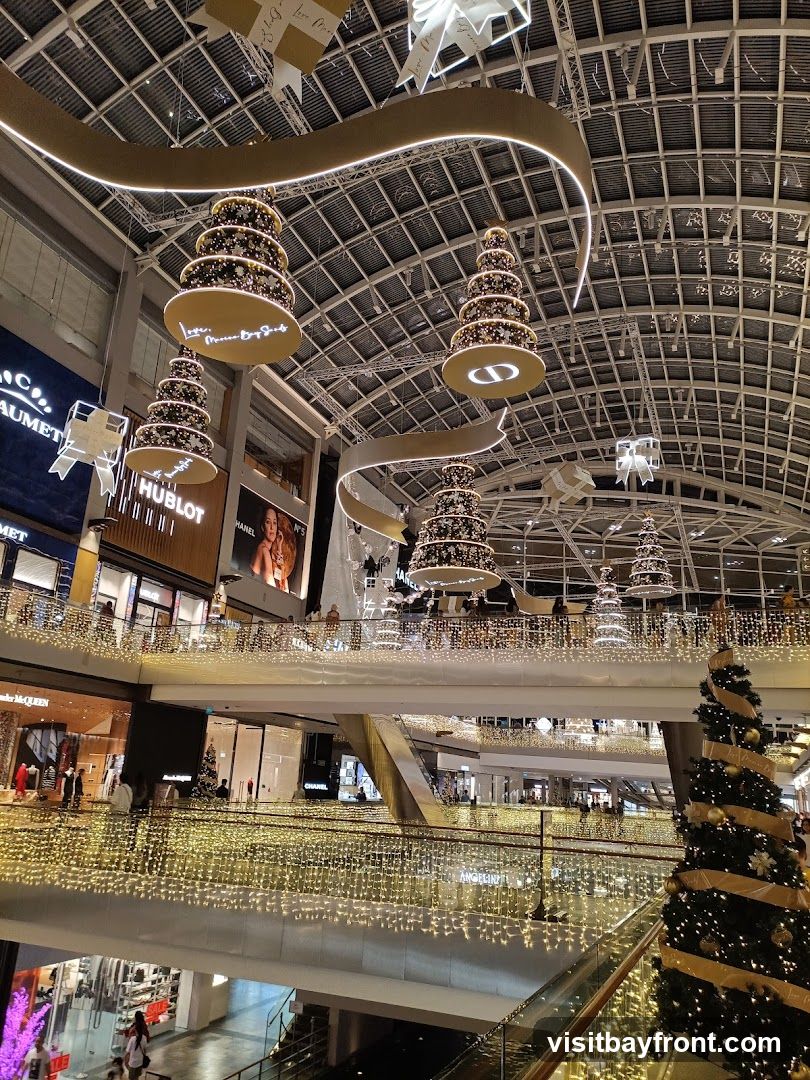 An interior view of a multi-level shopping mall decorated for Christmas. Large, tiered Christmas tree ornaments hang from the ceiling, adorned with lights and gold ribbons. The mall's interior is illuminated by numerous strings of warm white fairy lights draped along the railings and balconies of each level. Storefronts with visible brand names like HUBLOT, CHANEL, and ALEXANDER MCQUEEN are present on the lower levels. A large Christmas tree stands on the right side of the image.