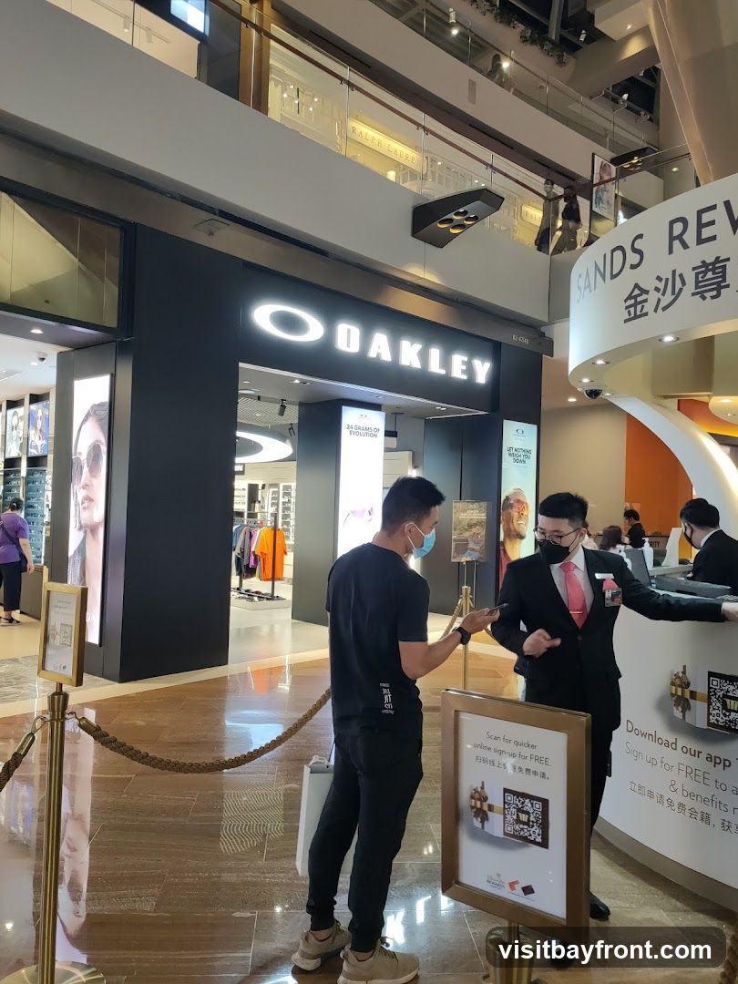 The exterior of an Oakley store is visible, with the brand's logo prominently displayed. Two men are interacting near a sign with a QR code. In the background, other stores and shoppers can be seen on different levels of the mall.