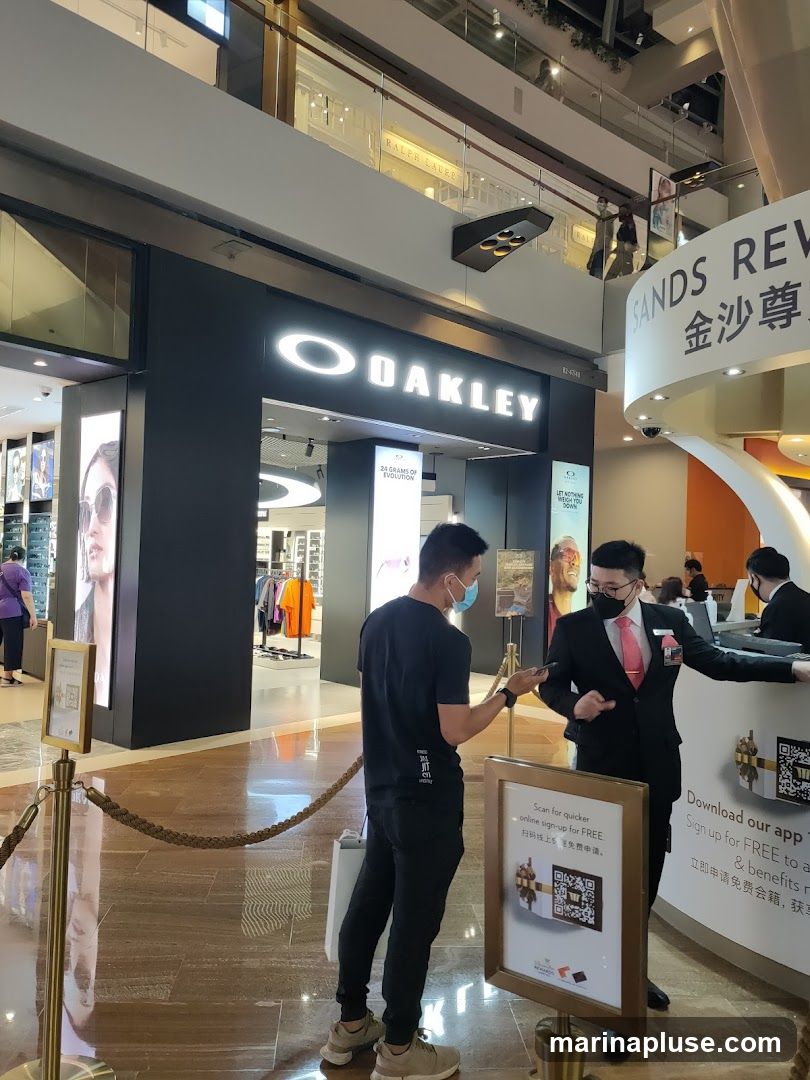 The exterior of an Oakley store is visible, with the brand's logo prominently displayed. Two men are interacting near a sign with a QR code. In the background, other stores and shoppers can be seen on different levels of the mall.