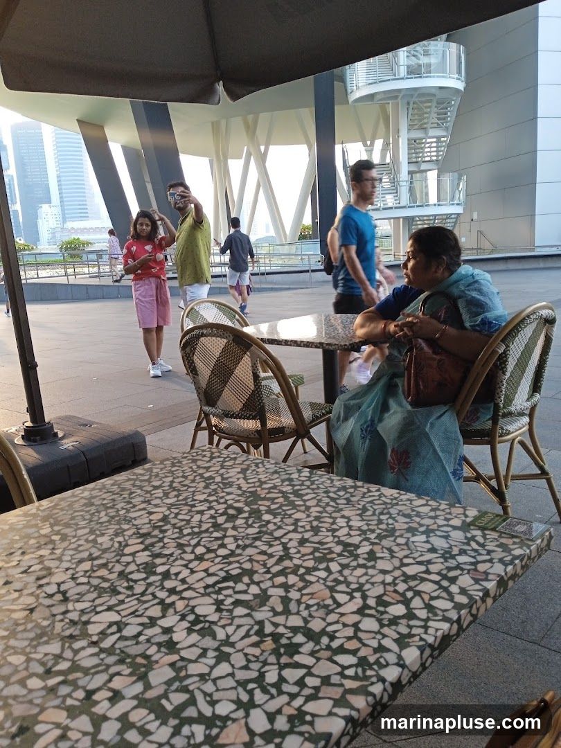 An outdoor seating area with tables and chairs, likely a cafe or restaurant. People are seen walking and sitting in the background. The architecture in the background suggests a modern urban setting. The tables have a terrazzo-like pattern.