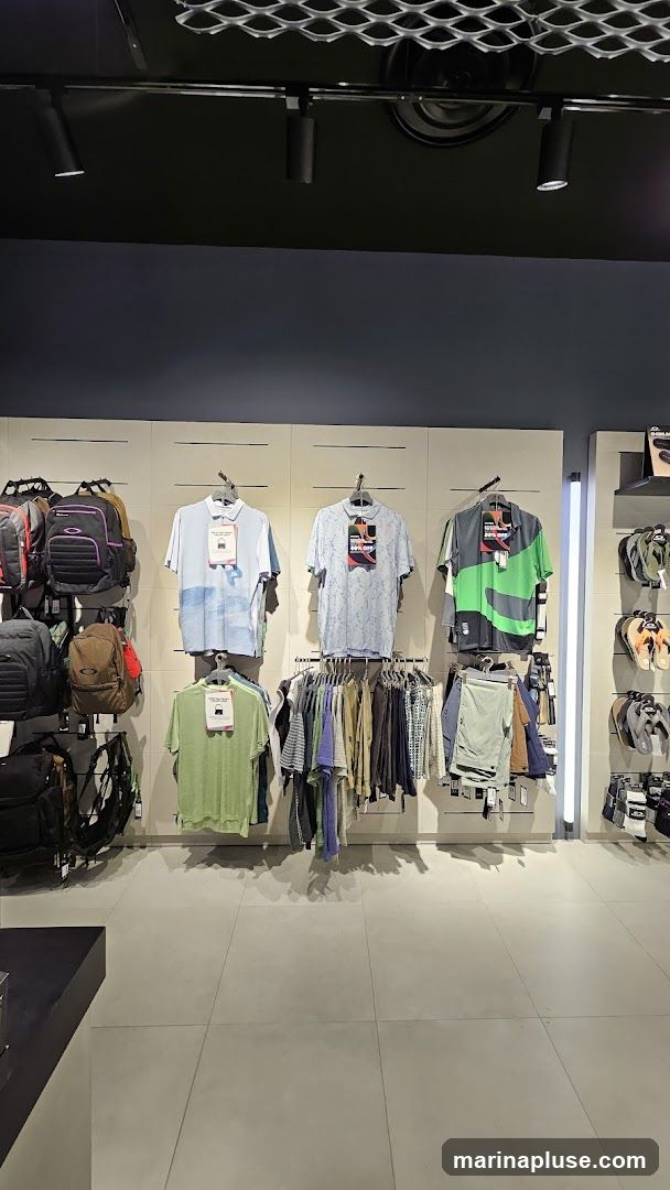 A retail display featuring various clothing items and accessories. On the left, several backpacks are hung on a wall. In the center, polo shirts and t-shirts are displayed on hangers, with some featuring sale tags. Below these, a rack holds a variety of folded shirts and shorts. To the right, more apparel is visible, along with a display of flip-flops and socks.
