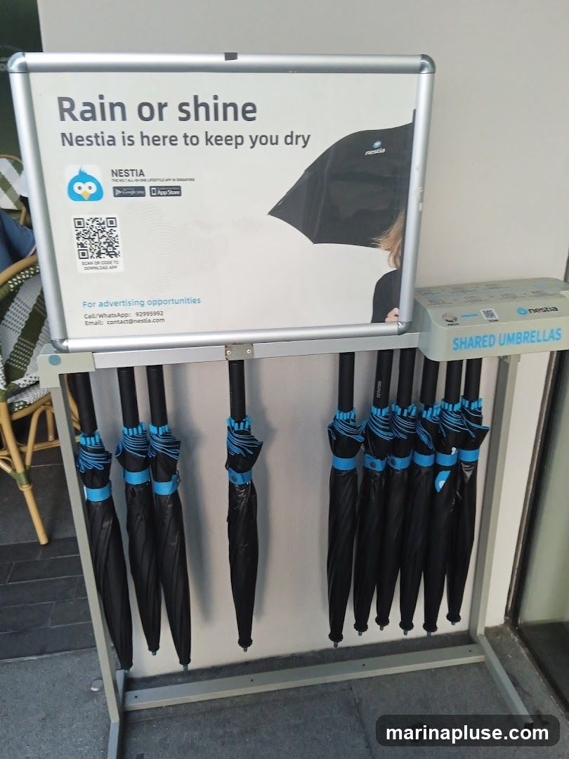 A rack of black umbrellas with blue accents is displayed under a sign that reads "Rain or shine Nestia is here to keep you dry". The sign also features a QR code for downloading the Nestia app and contact information for advertising opportunities. Below the main sign, a smaller sign reads "SHARED UMBRELLAS" and also displays the Nestia logo.