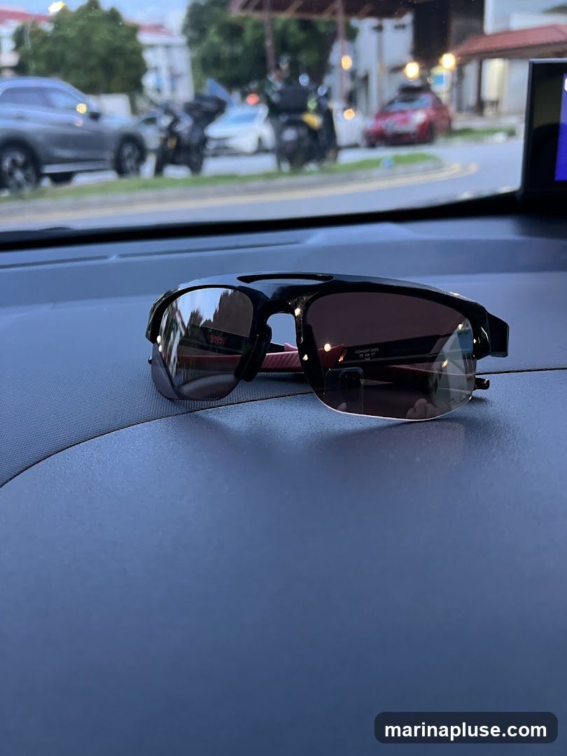 A pair of black sports sunglasses with tinted lenses rests on a dark blue dashboard. The background is blurred, showing a street scene with parked cars and motorcycles, and buildings under a twilight sky.