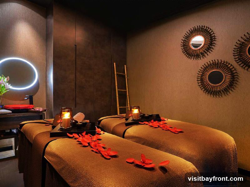 A dimly lit spa room with two massage beds, each adorned with red flower petals and a lit candle in a glass holder. A circular mirror with a neon light surrounds it is visible on the left, along with a sink and some flowers. Three decorative sunburst mirrors hang on the textured wall on the right. A bamboo ladder is placed between the beds and the wall.