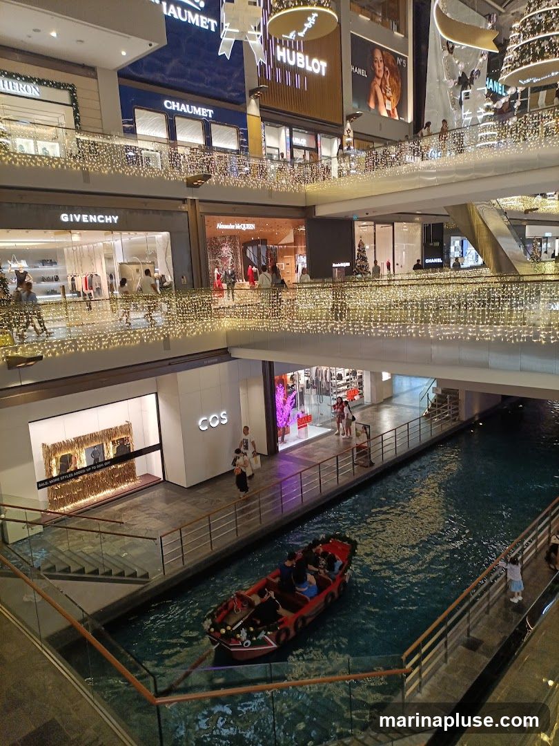An indoor shopping mall with multiple levels, decorated with numerous string lights. A canal runs through the lower level with a boat carrying passengers. Various luxury brand stores are visible on the upper levels, including Givenchy, Alexander McQueen, COS, Chaumet, and Hublot.