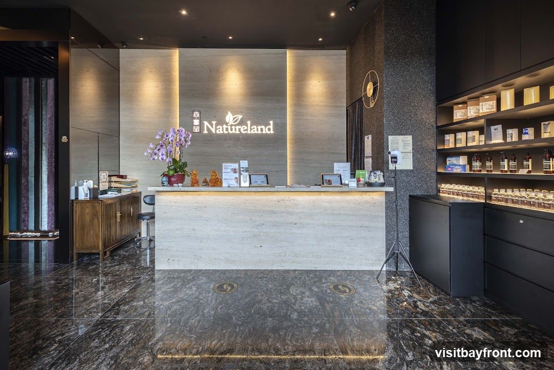 A well-lit reception area with a prominent "Natureland" logo on a marble-clad wall. The reception desk is made of light-colored marble and features several digital displays and decorative items, including a potted orchid. To the left, a wooden cabinet holds various items. To the right, shelves display numerous bottles and boxes of products. The floor is made of dark, polished stone.