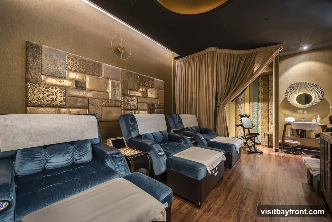 A room with several plush, teal-colored massage chairs arranged in a row. The chairs have footrests and are covered with white protective sheets. Behind the chairs is a textured wall with a decorative panel featuring gold accents. To the right, a massage chair and a sink area are visible, separated by curtains. The floor is made of dark wood.