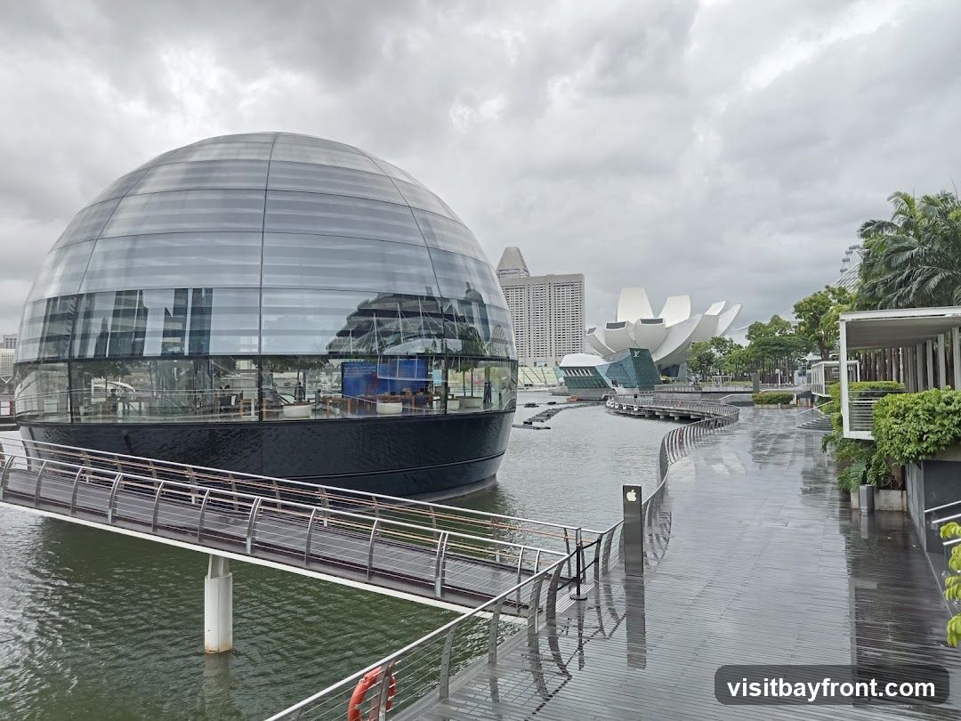 A view of the Apple Store at Marina Bay Sands in Singapore on a cloudy day. The store is a glass dome structure situated on the water, with a walkway leading to it. In the background, other buildings and the ArtScience Museum are visible.