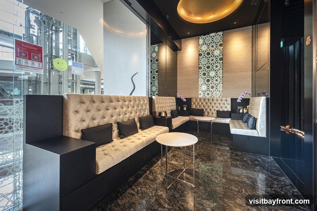 A sophisticated seating area with plush, tufted banquettes in a neutral tone, accented by dark wood paneling and black throw pillows. Small, round marble-topped tables with chrome bases are placed strategically. The walls feature textured stone and intricate geometric patterned panels, illuminated by warm lighting. A golden, domed ceiling element adds a touch of opulence. In the background, through a glass partition, a modern shopping mall interior with escalators and retail displays is visible. A few small orchid arrangements add a delicate floral touch.