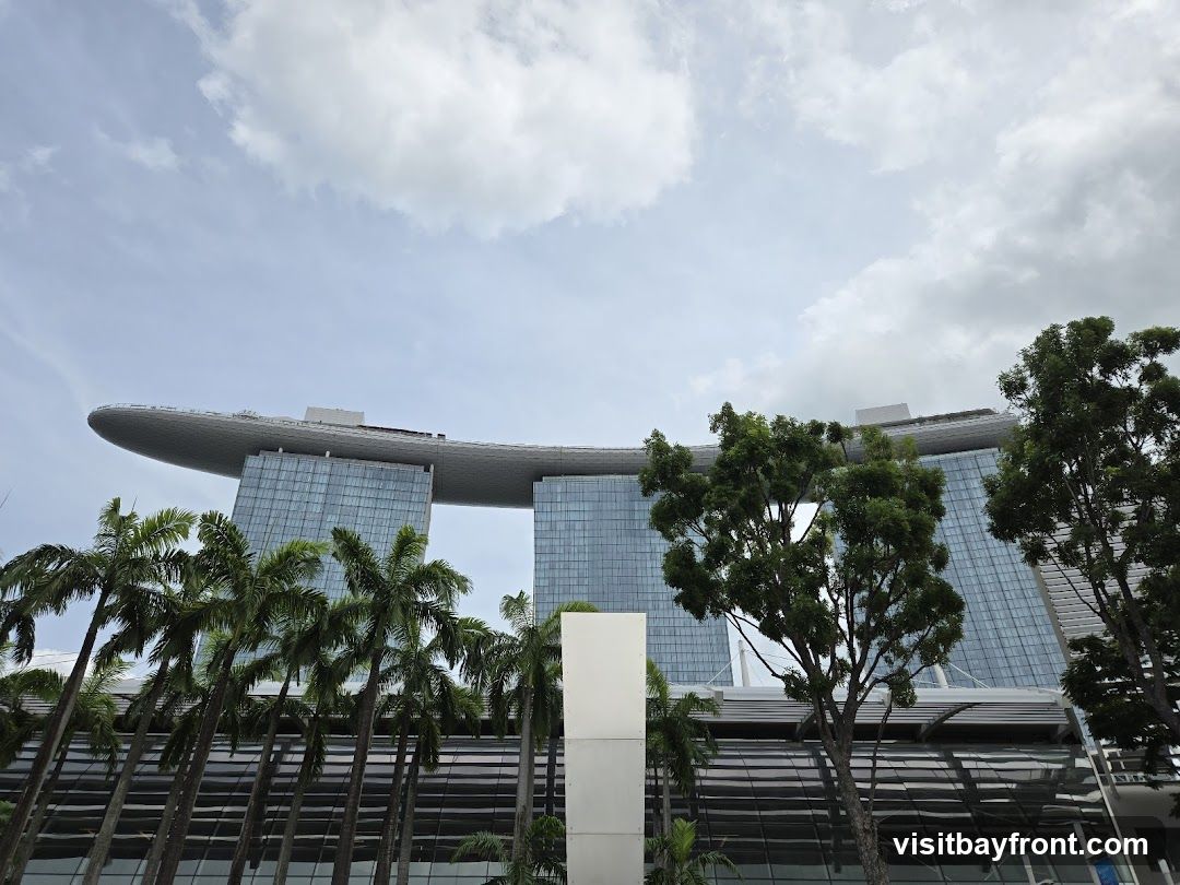 A low-angle shot of the Marina Bay Sands hotel in Singapore, with palm trees in the foreground. The sky is cloudy.