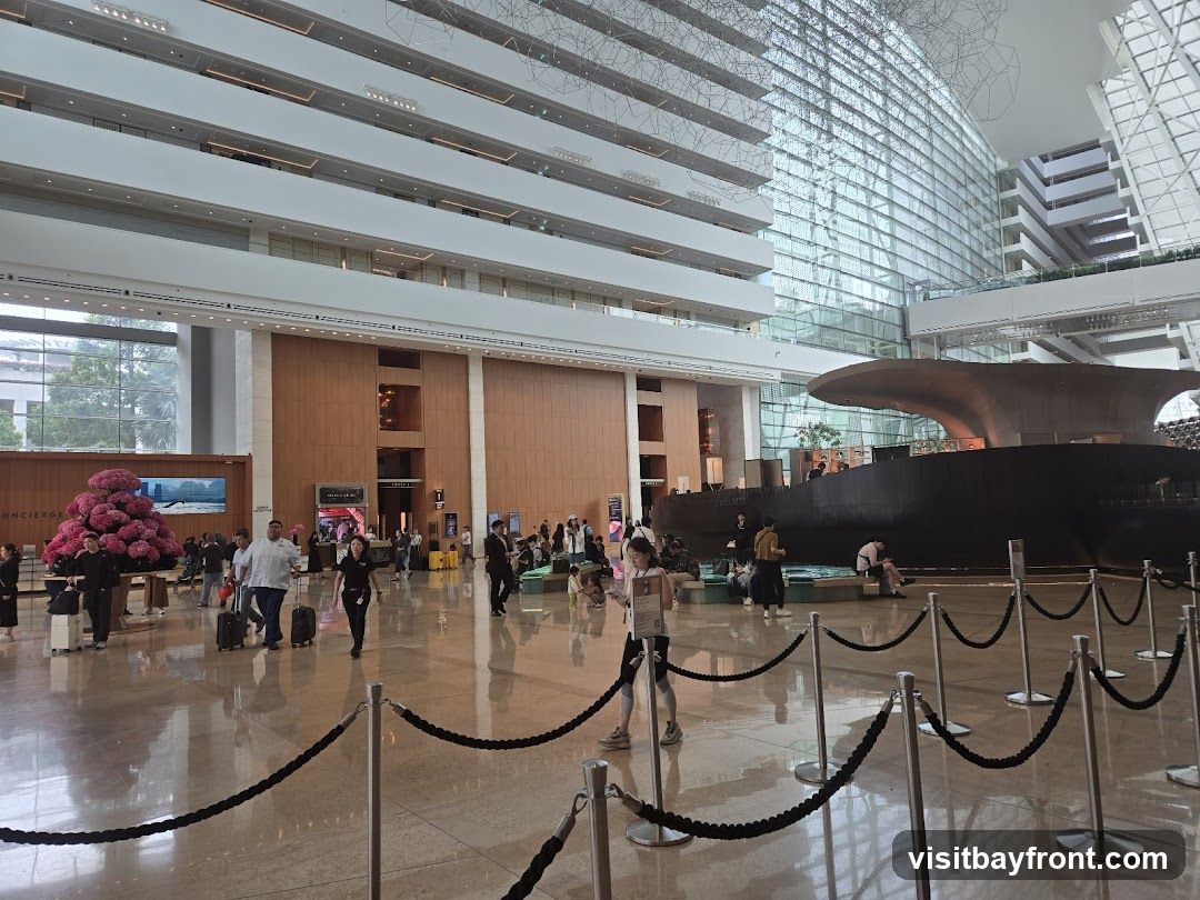 A wide shot of a grand hotel lobby with a high ceiling and modern architecture. People are seen walking with luggage, and there are various seating areas. A large, dark, boat-like sculpture is a prominent feature in the center of the space. The lobby has a polished floor reflecting the lights and the surrounding structures.
