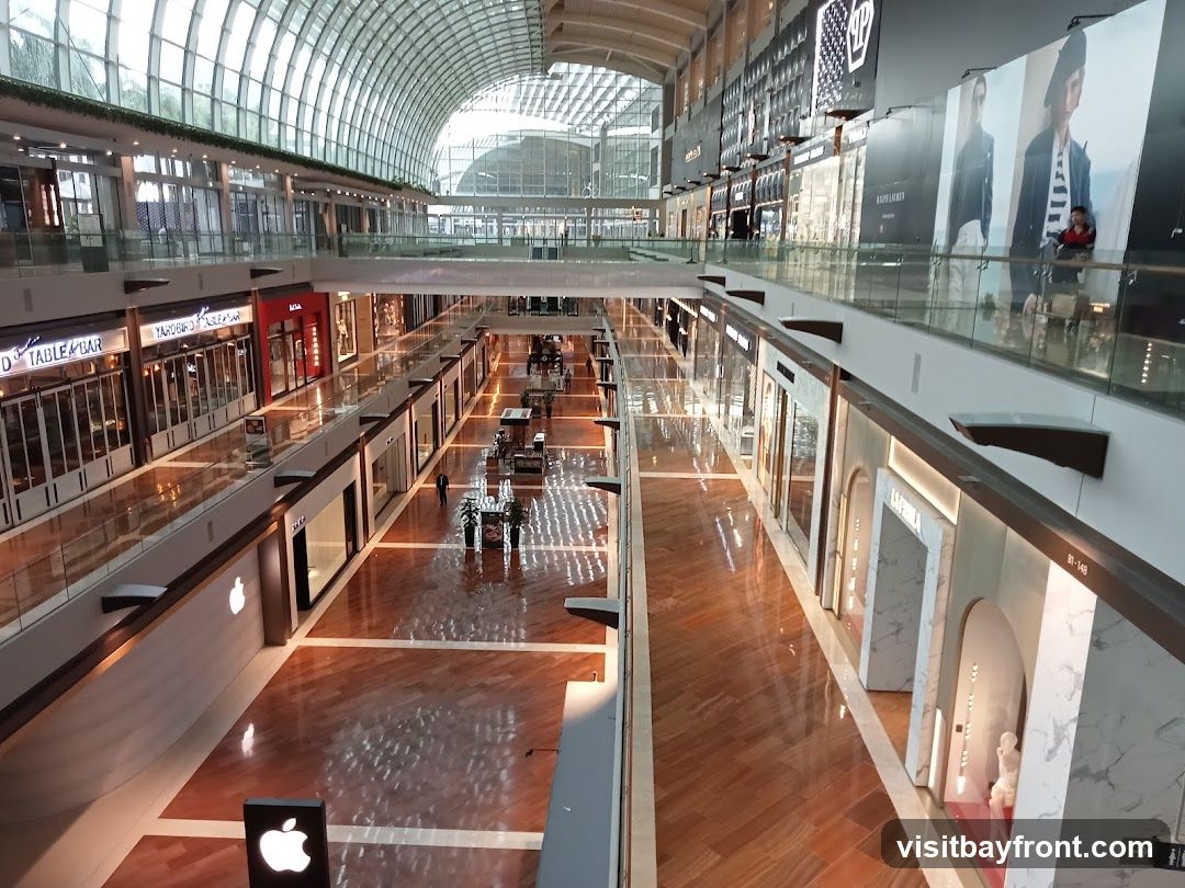 A wide-angle view of the interior of a large, modern shopping mall. The mall features multiple levels with shops lining the walkways. The architecture includes a distinctive arched glass roof that allows natural light to filter in. The floors are made of polished wood, reflecting the lights and storefronts. Several shops are visible, including one with a prominent Apple logo. Large advertisements and displays are mounted on the walls of the upper levels. People can be seen walking through the mall.