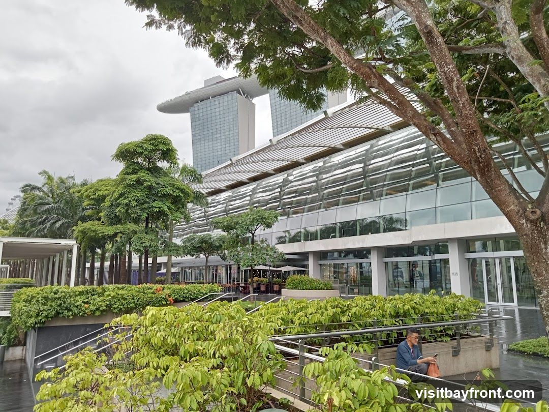 A wide shot of a modern building with a curved glass facade, set against a cloudy sky. Lush green plants and trees surround the building, with a man sitting on a ledge in the foreground. In the background, the iconic Marina Bay Sands hotel is visible.