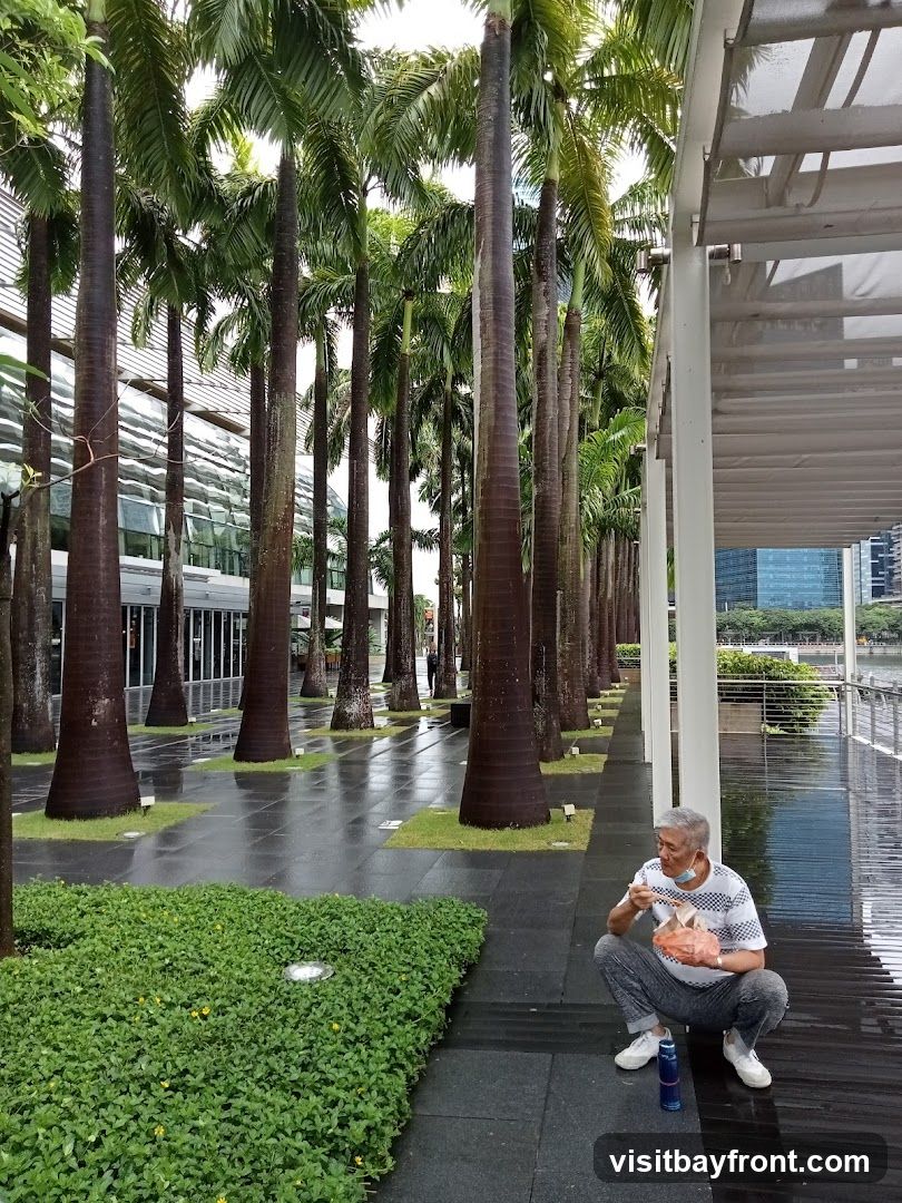 An elderly Asian man wearing a face mask and a checkered shirt squats on a wooden walkway, eating from a takeout container with chopsticks. He is surrounded by tall palm trees and lush green foliage. The ground is wet, suggesting recent rain. In the background, a modern building with large glass windows is visible.