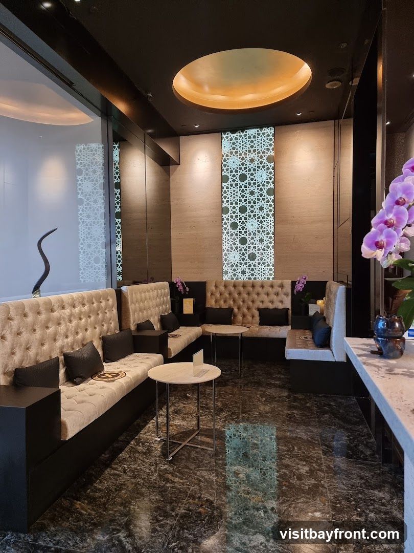 A sophisticated lounge area with plush, tufted banquette seating in a neutral tone, accented with dark throw pillows. Small, round marble-topped tables are placed between the seating sections. The walls feature a textured stone finish and decorative geometric panels that are illuminated from behind. A large, circular ceiling fixture with warm lighting adds to the ambiance. A vase with purple orchids is visible on the right side of the image, near a counter. The floor is a dark, polished marble with a marbled pattern.