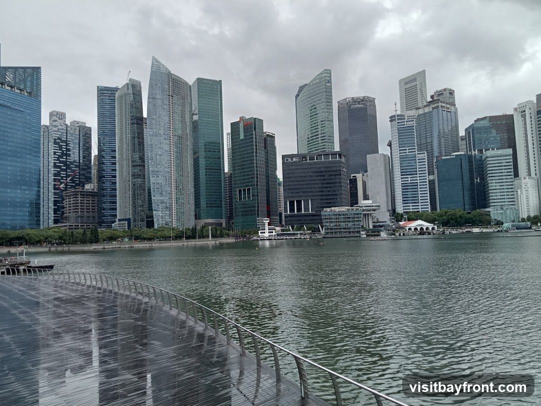 A panoramic view of a modern city skyline with numerous skyscrapers of varying heights and architectural styles, situated along a wide body of water. The sky is overcast with clouds. In the foreground, a wet, reflective walkway with a metal railing curves into the scene. The water appears calm with ripples. Several boats are visible on the water in the distance. The buildings are a mix of glass, steel, and concrete, with some featuring distinct shapes and designs. Signs for companies like OUE, Citi, and WeWork are visible on some of the buildings.