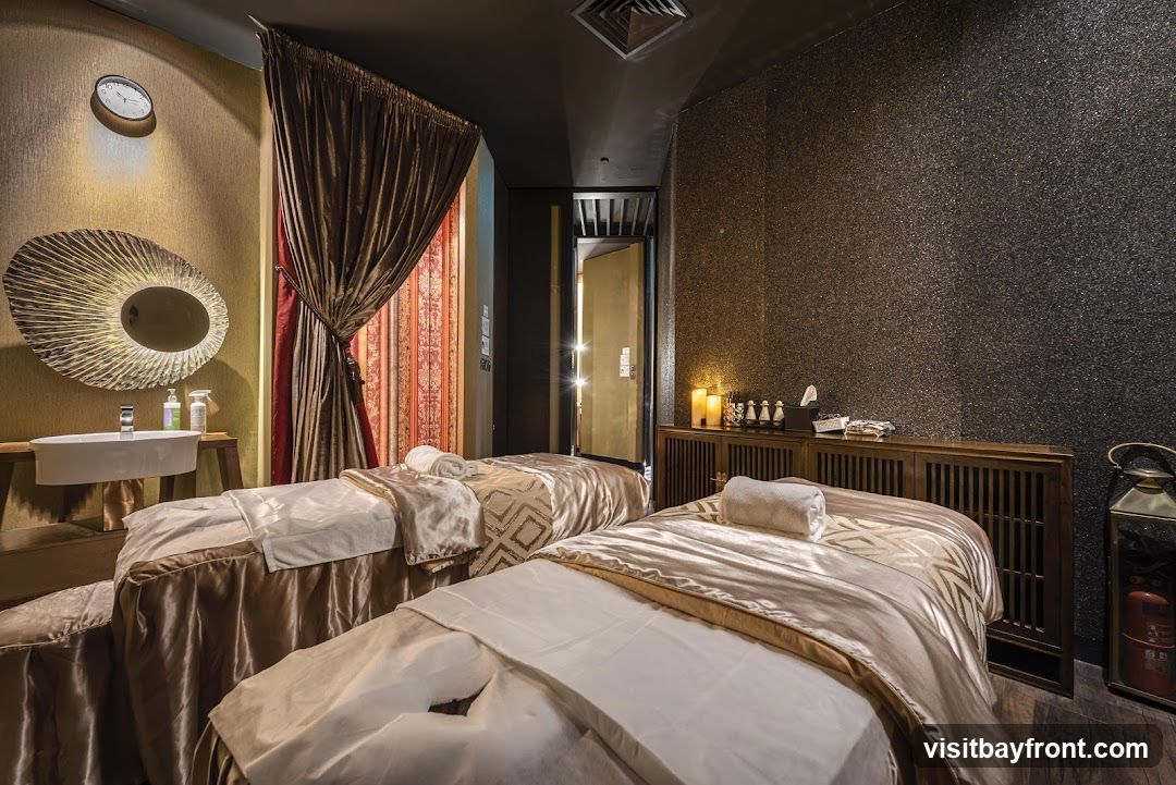 A luxurious spa treatment room with two massage beds, each covered in satin sheets and topped with a rolled towel. To the left, a sink is mounted on a wooden stand beneath a decorative mirror and a wall clock. Heavy drapes in shades of brown and red frame the beds. To the right, a wooden cabinet holds candles, bottles of oil, and other spa amenities. The walls are textured, one with a shimmering dark finish and the other with a golden hue.