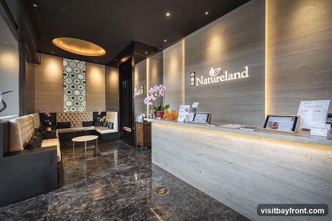 A modern and luxurious spa reception area with a marble counter, comfortable seating, and elegant decor. The Natureland logo is visible on the wall behind the counter. There are tablets on the counter displaying information and a sign indicating social distancing.