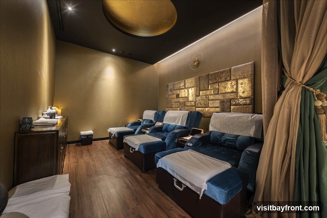 A spa room with multiple plush blue massage chairs lined up, each with a footrest. The walls are a textured gold, and the ceiling features a large, circular gold accent. A dark wooden cabinet on the left holds various spa products. Decorative curtains in green and beige hang on the right side of the room.