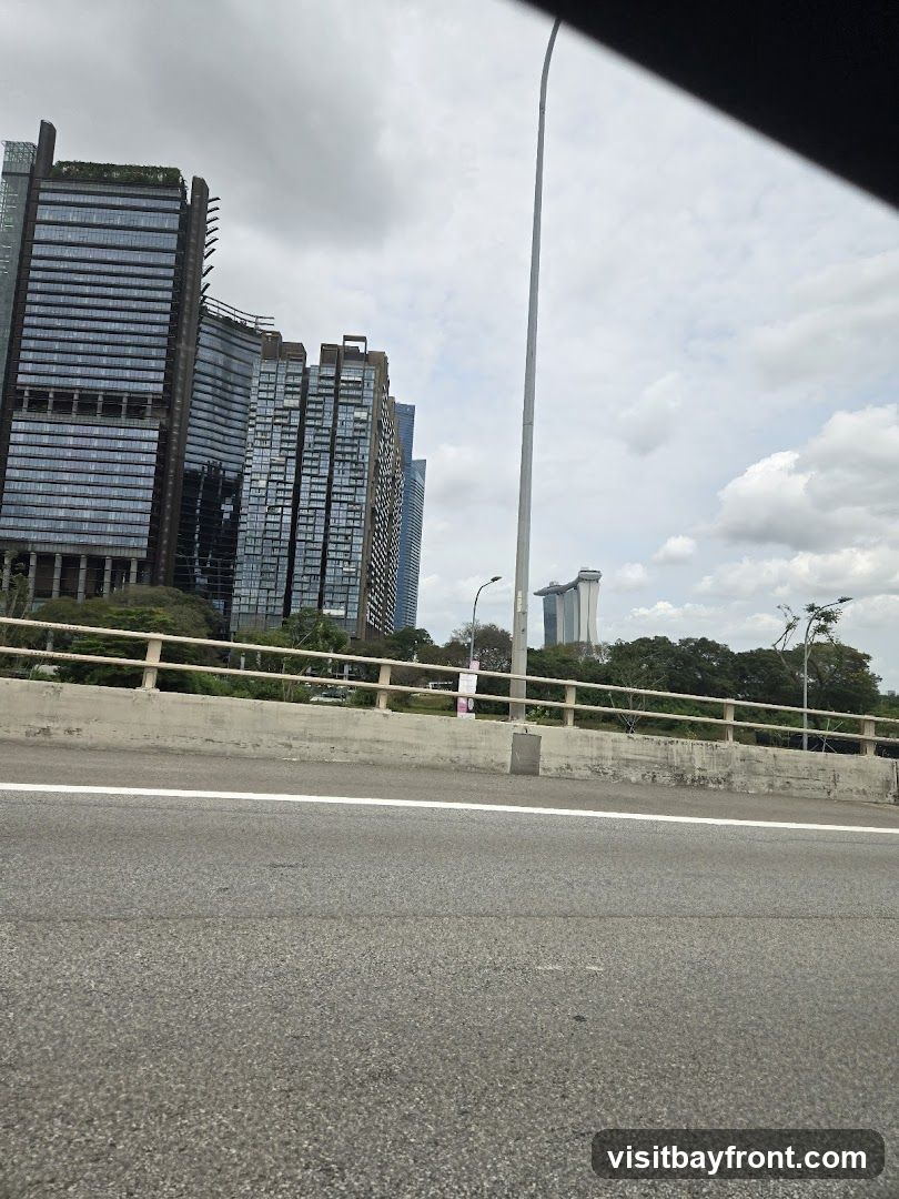 A view of a city skyline featuring several modern, tall buildings with glass facades. The buildings are clustered together, with some appearing to have green roofs or terraces. In the distance, another distinctive building with a unique architectural design is visible. The foreground shows a highway or elevated road with a concrete barrier and railing. The sky is overcast with scattered clouds.