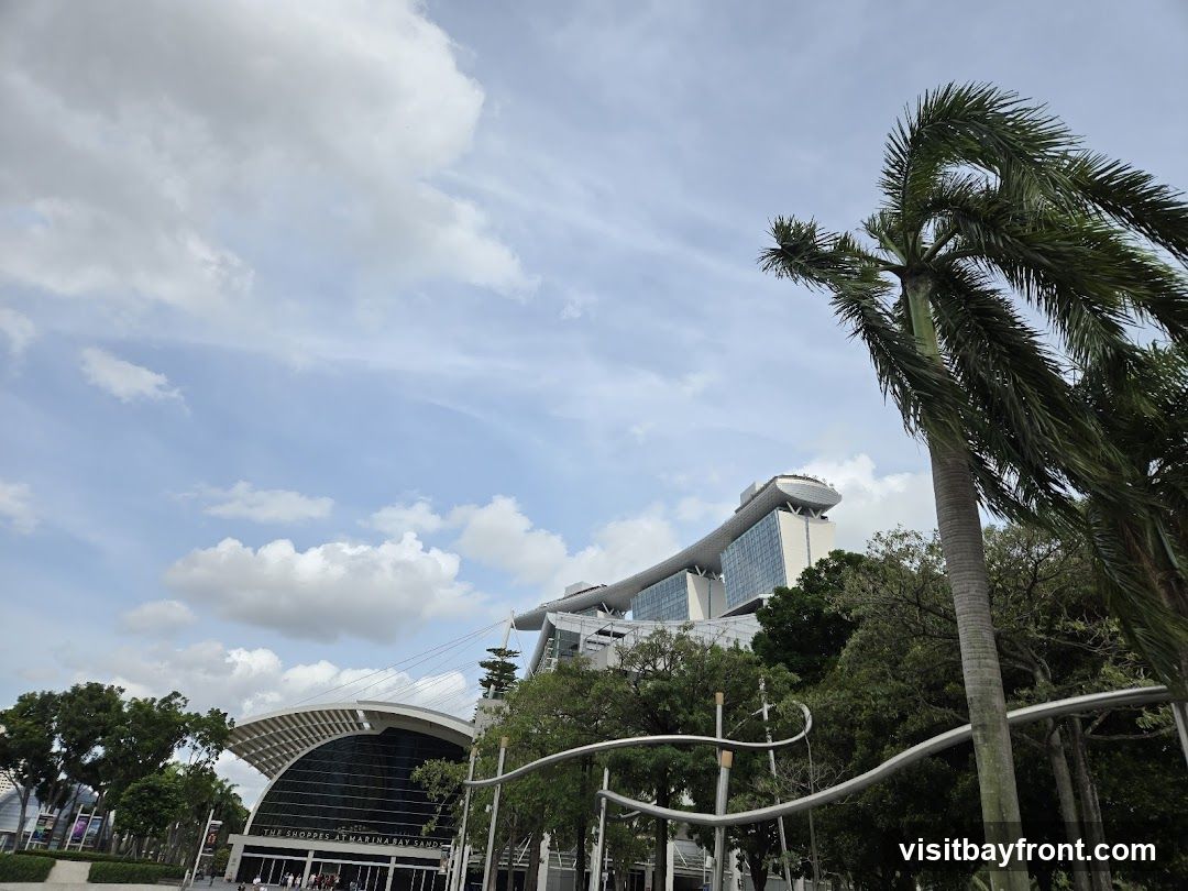 A wide-angle shot captures the striking modern architecture of The Shoppes at Marina Bay Sands, with its distinctive curved roof and glass facade, set against a bright blue sky dotted with fluffy white clouds. Lush green trees and a tall palm tree with fronds swaying in the breeze frame the scene, adding a tropical feel to the urban landscape.