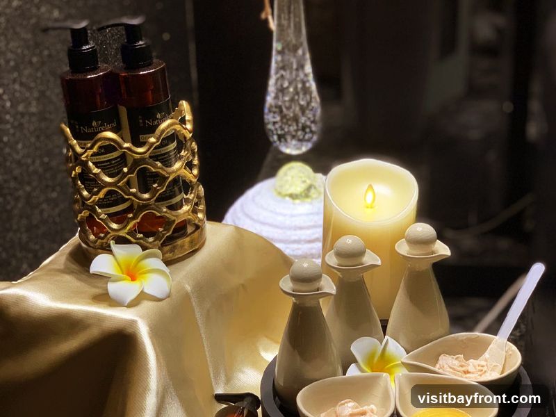 A close-up shot of spa products arranged on a golden cloth. Two dark bottles with pump dispensers, labeled 'Natureland Organic Oil', are held in a decorative gold stand. In front of the stand, a white plumeria flower rests on the cloth. To the right, a large LED candle flickers, casting a warm glow. Beside the candle are three small ceramic bottles with stoppers and several small bowls containing creams or balms, one with a spatula. A decorative glass vase is visible in the background.