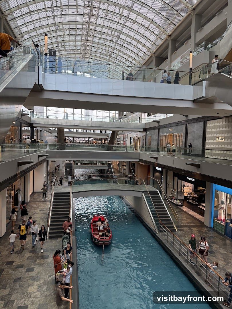 An interior view of a multi-level shopping mall with a canal running through the center. A red boat with passengers navigates the blue water. People are seen walking on the walkways and escalators on different levels. Shops with visible signage like 'CHLOE' and 'PAZZION' are present. The mall features a modern architectural design with a glass roof structure.