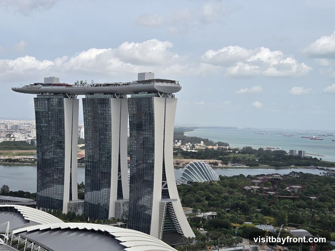 An aerial view of the Marina Bay Sands hotel in Singapore, with its iconic three towers topped by a ship-like structure. In the foreground, part of a stadium roof is visible. To the right, the Gardens by the Bay, including the Supertree Grove and the Cloud Forest and Flower Dome conservatories, are seen nestled amongst lush greenery. The Singapore Strait, dotted with numerous cargo ships, stretches across the horizon under a partly cloudy sky.