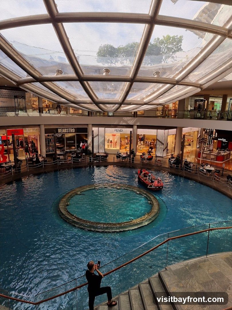An interior view of a modern shopping mall with a large, circular indoor water feature. A small boat with people is navigating the water. Shops with visible signage such as HUGO, BEANSTRO, and FENDI are located around the perimeter of the water feature. A person is standing on a staircase in the foreground, taking a photo with their phone. The mall has a large, geodesic dome-like glass roof.