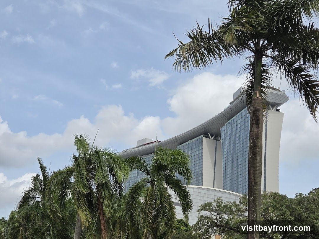 A view of a large, modern skyscraper with a distinctive curved roof structure, partially obscured by lush green palm trees and other foliage. The building features a facade of glass windows and white panels. The sky above is blue with scattered white clouds.