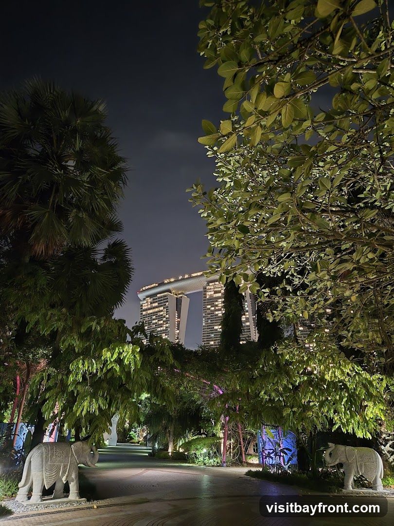 A paved pathway winds through a lush garden at night, illuminated by soft lights. Two stone elephant statues flank the entrance to the path. In the background, the iconic Marina Bay Sands hotel complex is visible, lit up against the dark sky. Trees with green foliage frame the scene.