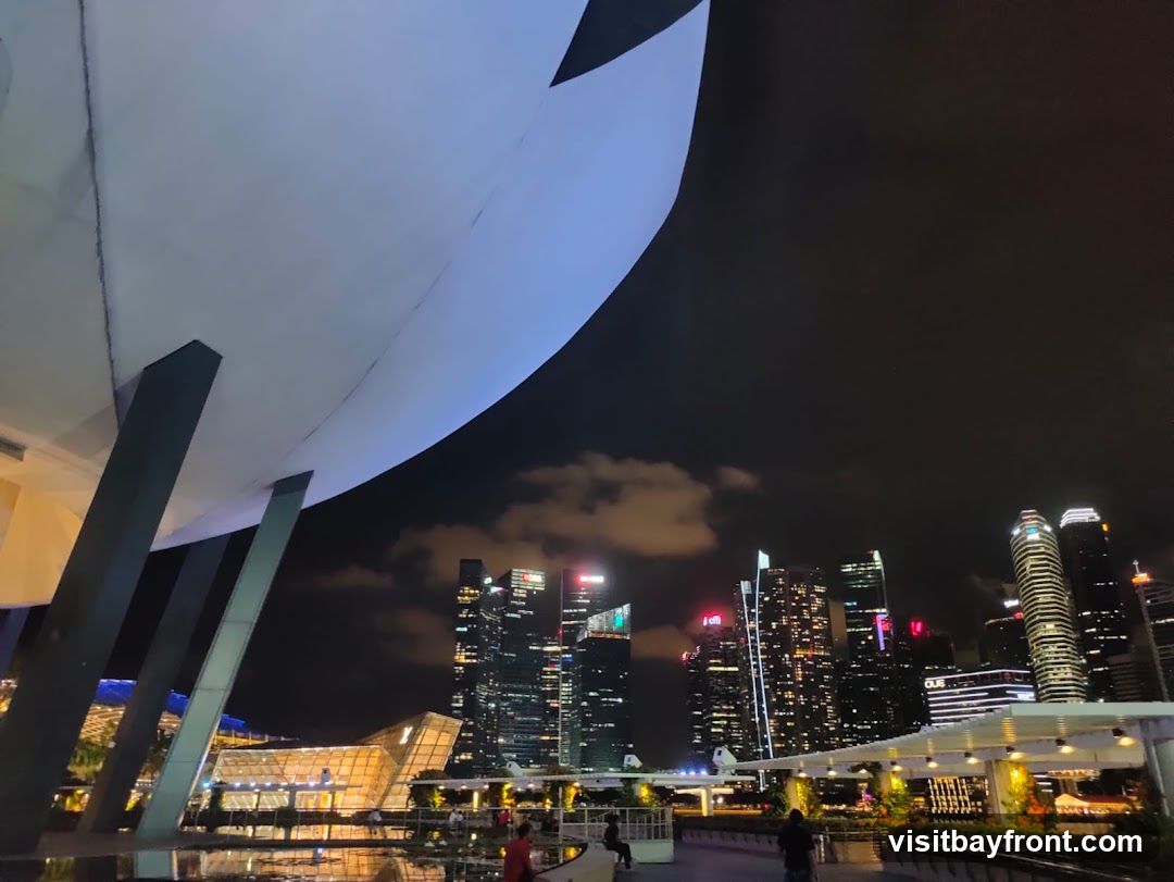 A wide-angle shot captures a city skyline at night, dominated by illuminated skyscrapers. In the foreground, a large, curved white structure supported by dark pillars creates a dramatic overhang. Below this structure, a reflective surface, possibly water or polished ground, mirrors the lights of the city. In the mid-ground, a modern building with a distinctive geometric design and a glass facade is visible, along with other structures and walkways. People can be seen strolling along the illuminated pathways, enjoying the urban nightscape. The sky is dark with scattered clouds, and the overall ambiance is one of a vibrant, modern metropolis.