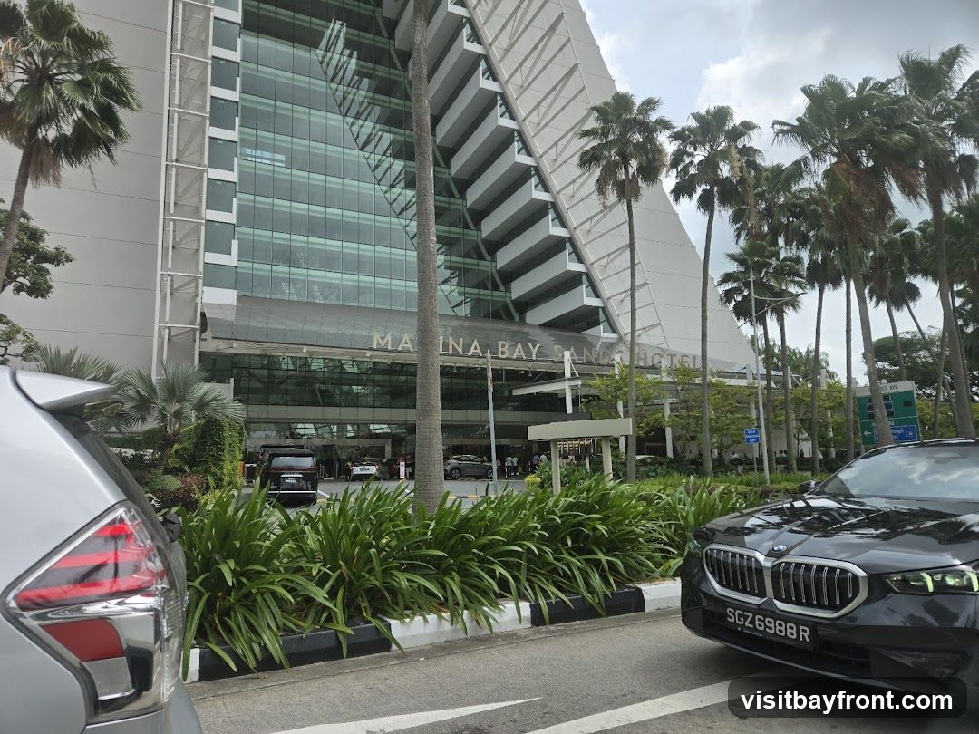 A view of the entrance to the Marina Bay Sands Hotel, with palm trees and cars in the foreground. The hotel's name is visible above the entrance.