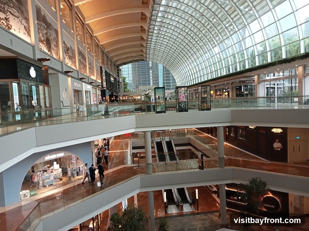 A wide-angle view of the interior of a multi-level shopping mall with a distinctive arched glass roof. The mall features escalators, walkways, and various storefronts. Large decorative murals are visible on the upper walls. Natural light streams in through the glass ceiling, illuminating the polished floors and modern architecture. Several shops are visible, including one with the sign 'AIMER KIDS'.