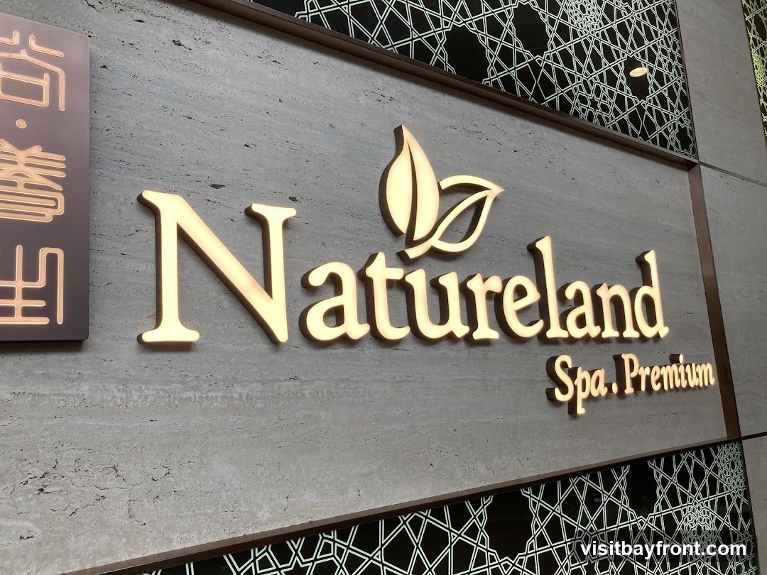A close-up shot of a sign for "Natureland Spa. Premium" mounted on a textured wall. The sign features illuminated golden lettering and a leaf-like logo. To the left, there is a separate sign with illuminated Chinese characters. The wall has a marble-like texture and is adorned with a decorative geometric pattern above and below the main sign.