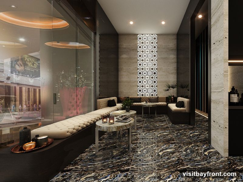 A sophisticated lounge area with plush seating, marble-patterned flooring, and decorative wall accents. A large window offers a view of a bustling interior space with a prominent "Into the Wild" advertisement. The ambiance is luxurious and inviting, with subtle lighting and decorative candles.