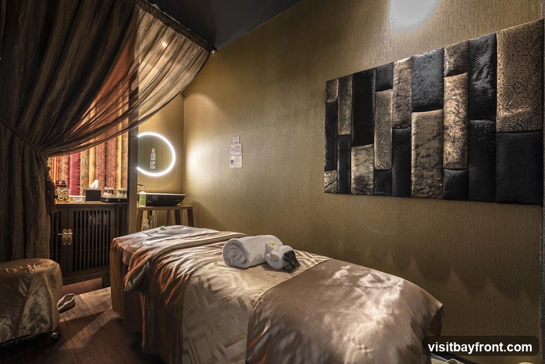 A massage room with a massage bed covered in satin sheets, towels, and a small decorative item. The room has gold textured walls, a dark wood cabinet with patterned curtains, a sink with a circular illuminated mirror, and a decorative wall panel.