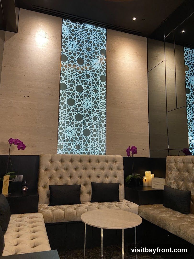 A luxurious lounge seating area with tufted velvet banquettes, black accent pillows, and a small round marble-top table. The wall features a decorative illuminated panel with an intricate geometric pattern, complemented by textured stone panels and mirrored surfaces. Two purple orchids add a touch of color, and LED candles provide ambient lighting.